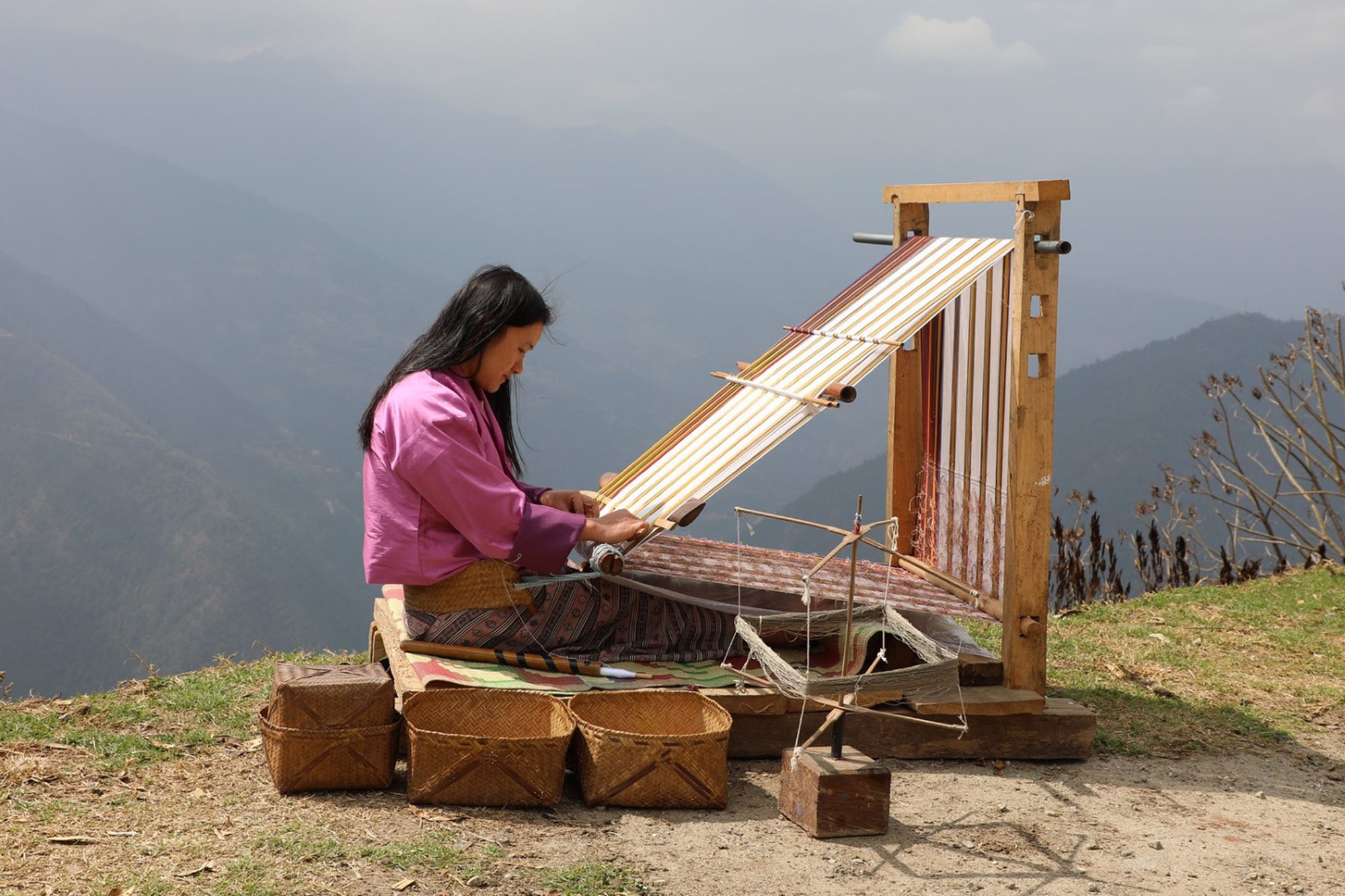 A woman seated outdoors on a grassy area near a mountainous landscape, operating a traditional wooden loom.