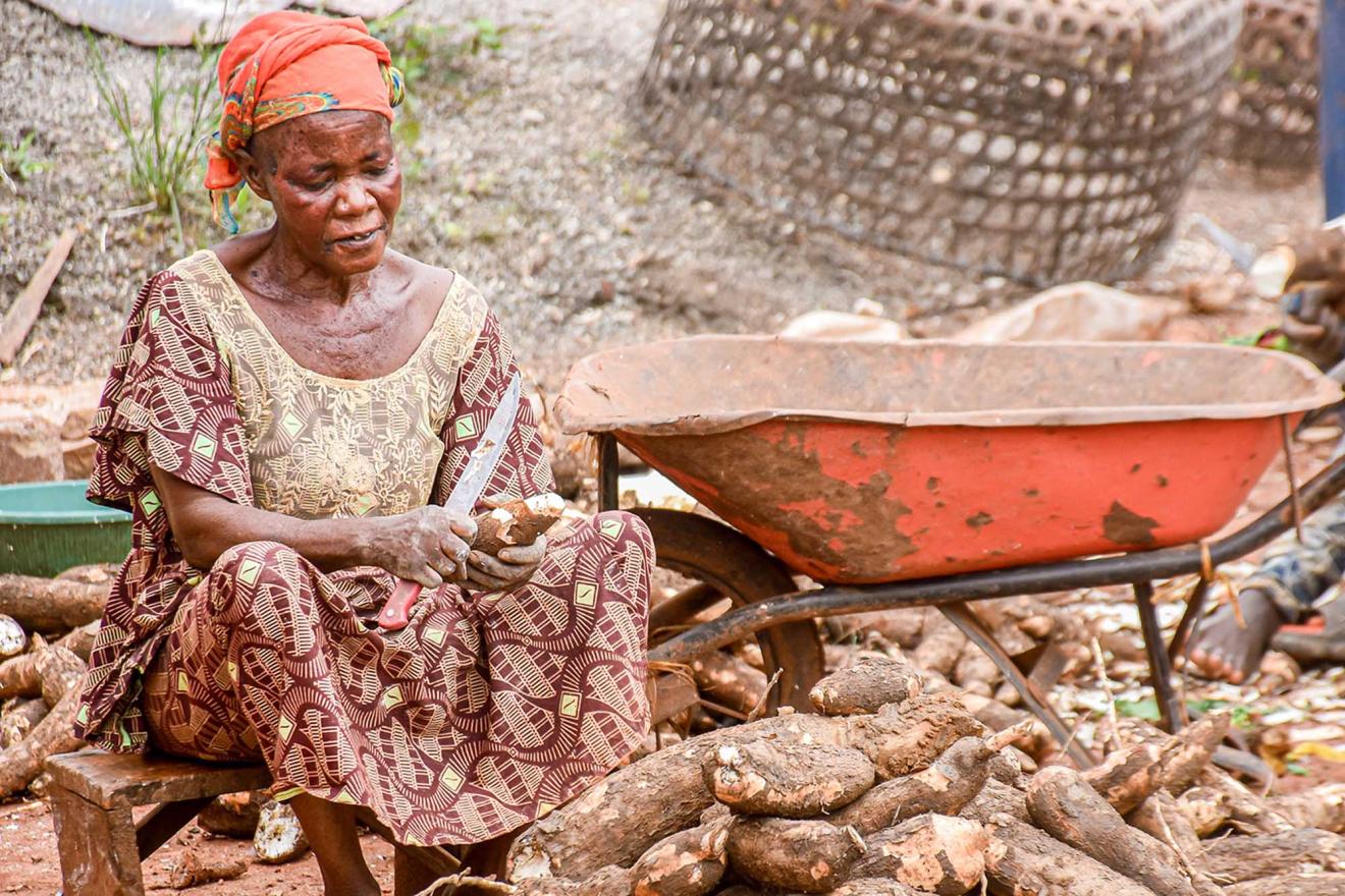 A woman sitting outdoors on a low stool, engaged in peeling or cutting tubers, which are scattered on the ground around them.