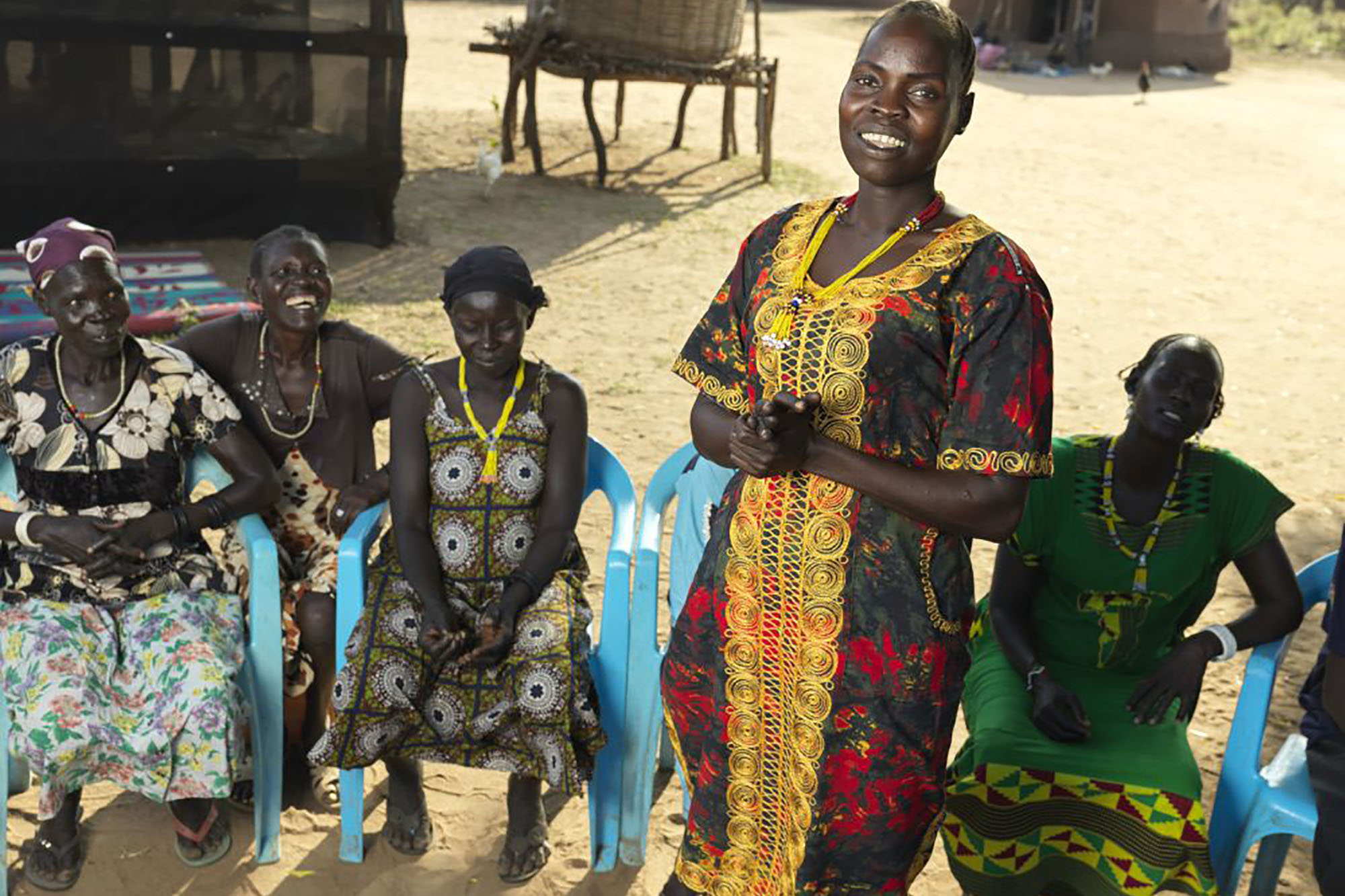Five women outdoors. Four of them are seated on blue plastic chairs while one stands in front of them.
