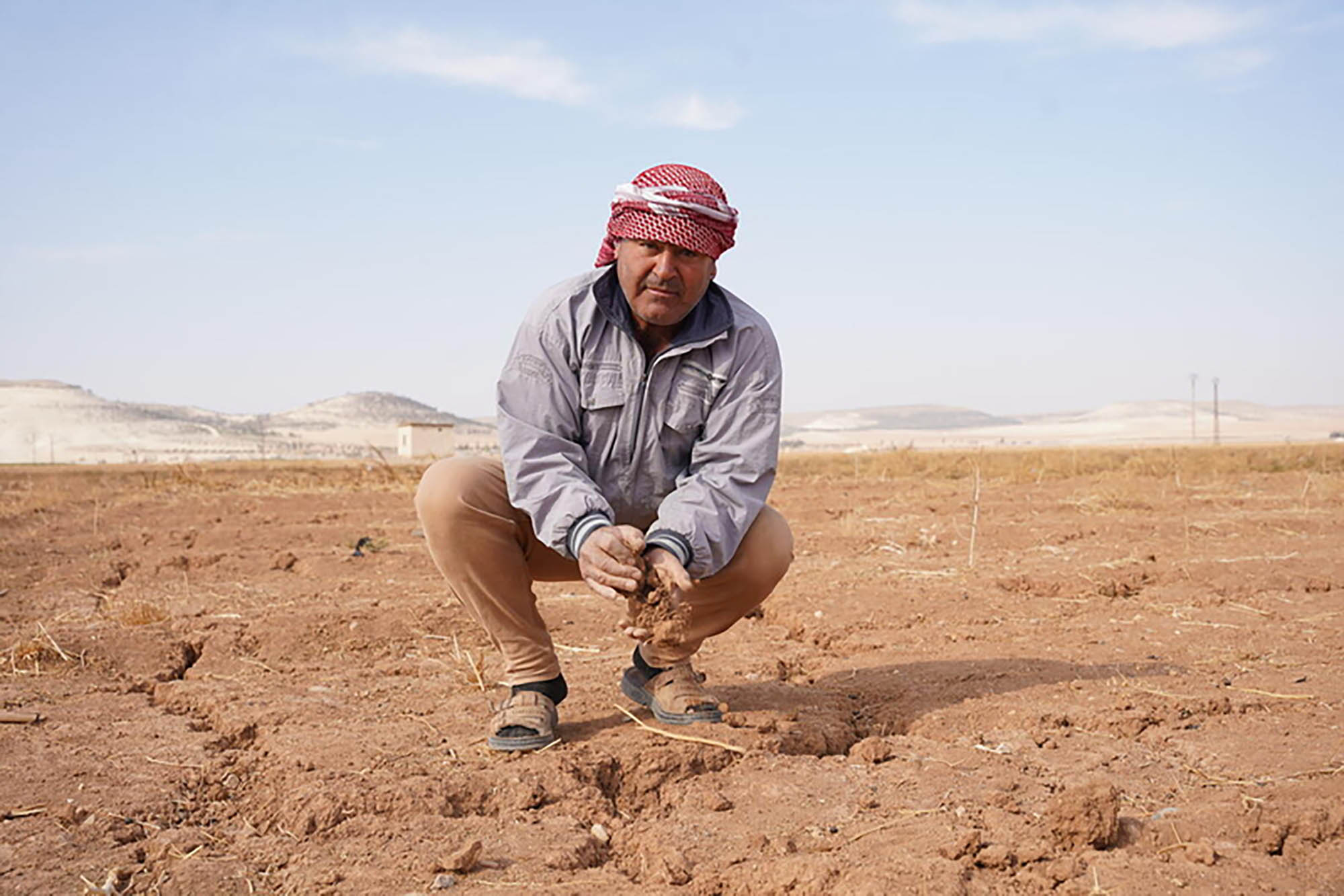 A man wearing a grey jacket, tan trousers and dark sandals is squatting on dry, cracked soil in an open field.