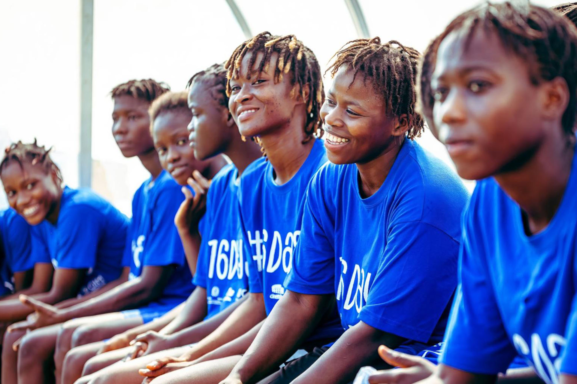 A group of female football players sitting in a row, wearing matching bright blue T-shirts. 