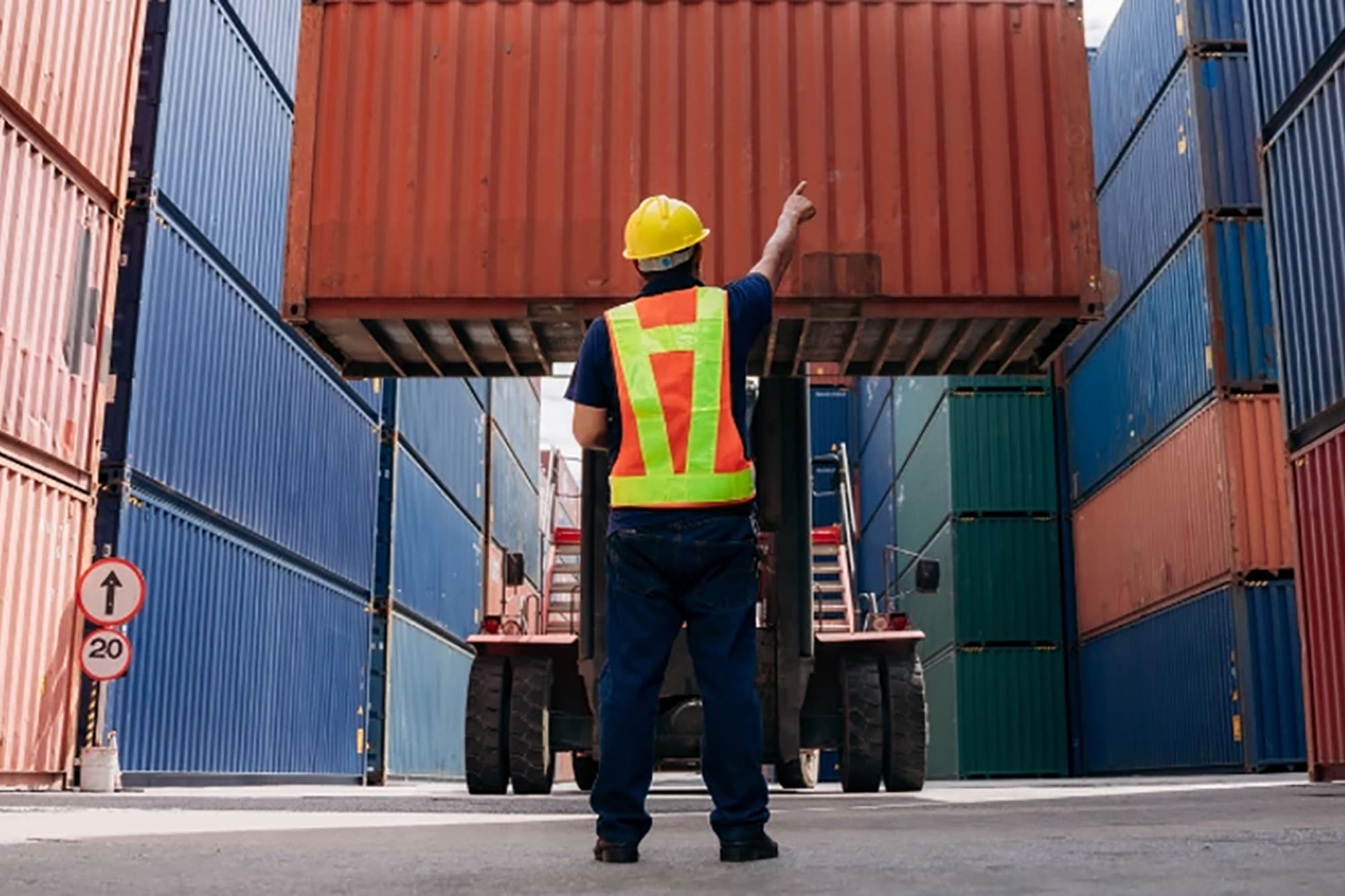 A worker wearing a yellow hard hat and a high-visibility safety vest is standing and pointing upward, guiding the placement of a container.