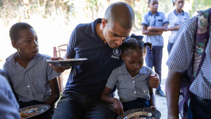 Jean-Martin leans sideways next to a girl in school uniform while they both are seated and eating from separate dishes