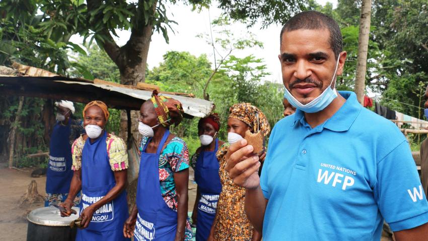Jean-Martin sampling a snack and his face-mask is on his chin while women are working in the background seen wearing theirs