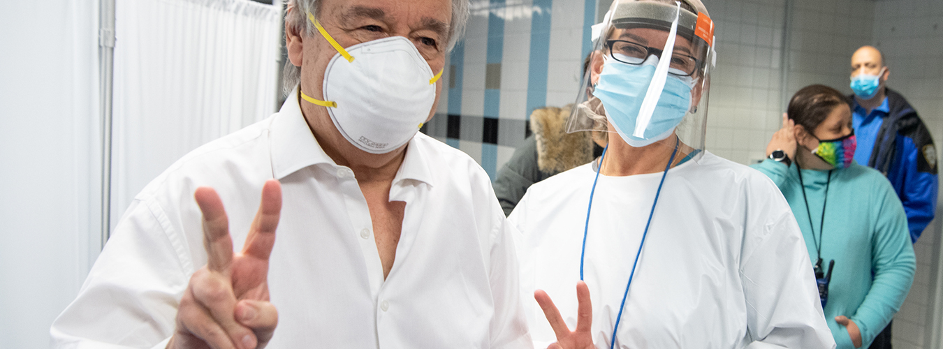 Secretary-General António Guterres and a nurse are wearing masks and both give the victory sign inside a medical setting.