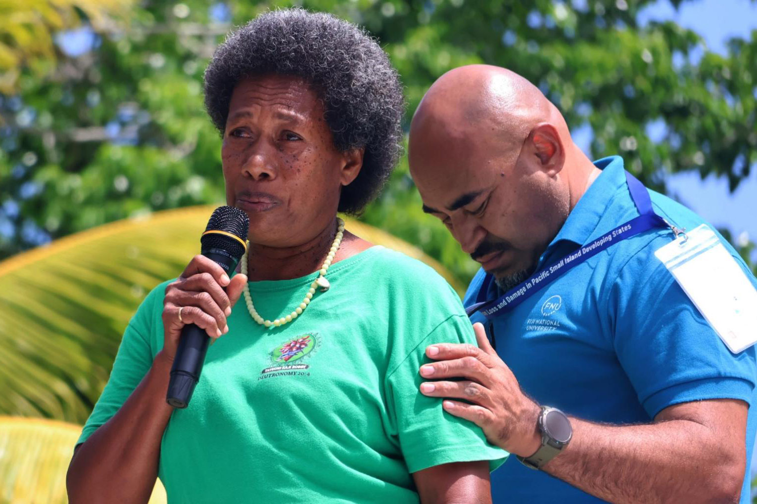 A woman holding a microphone with her colleague holding her shoulder.
