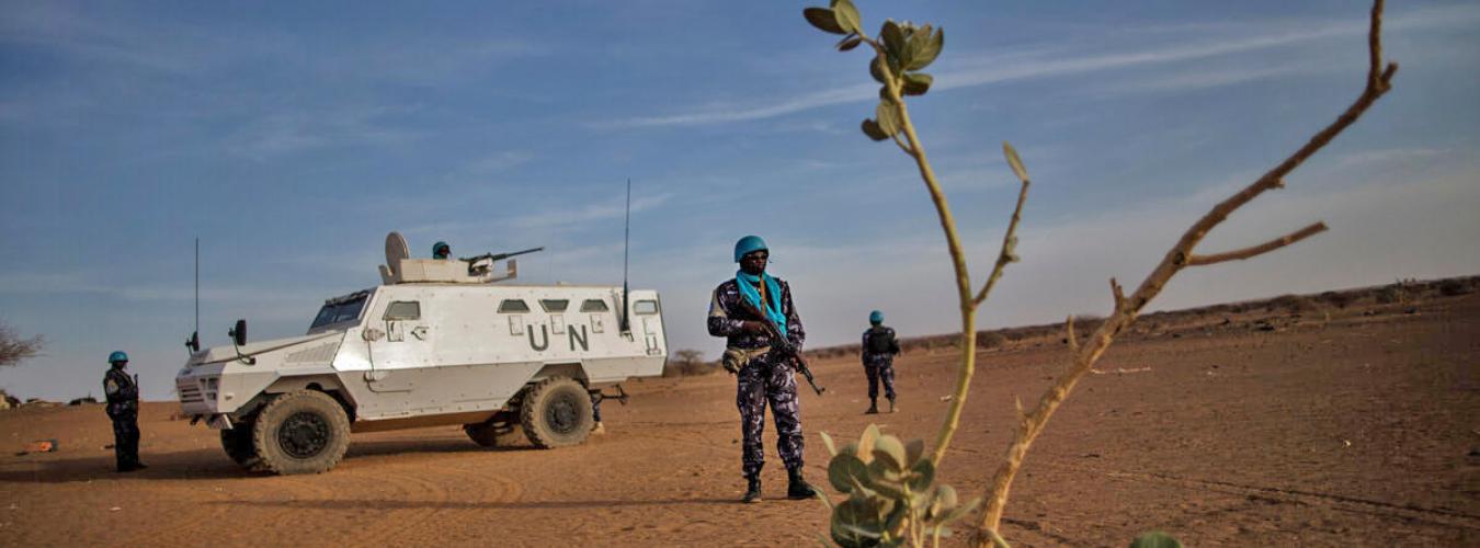 A UN peacekeeper standing guard with 2 peacekeepers and a UN vehicle in the background.