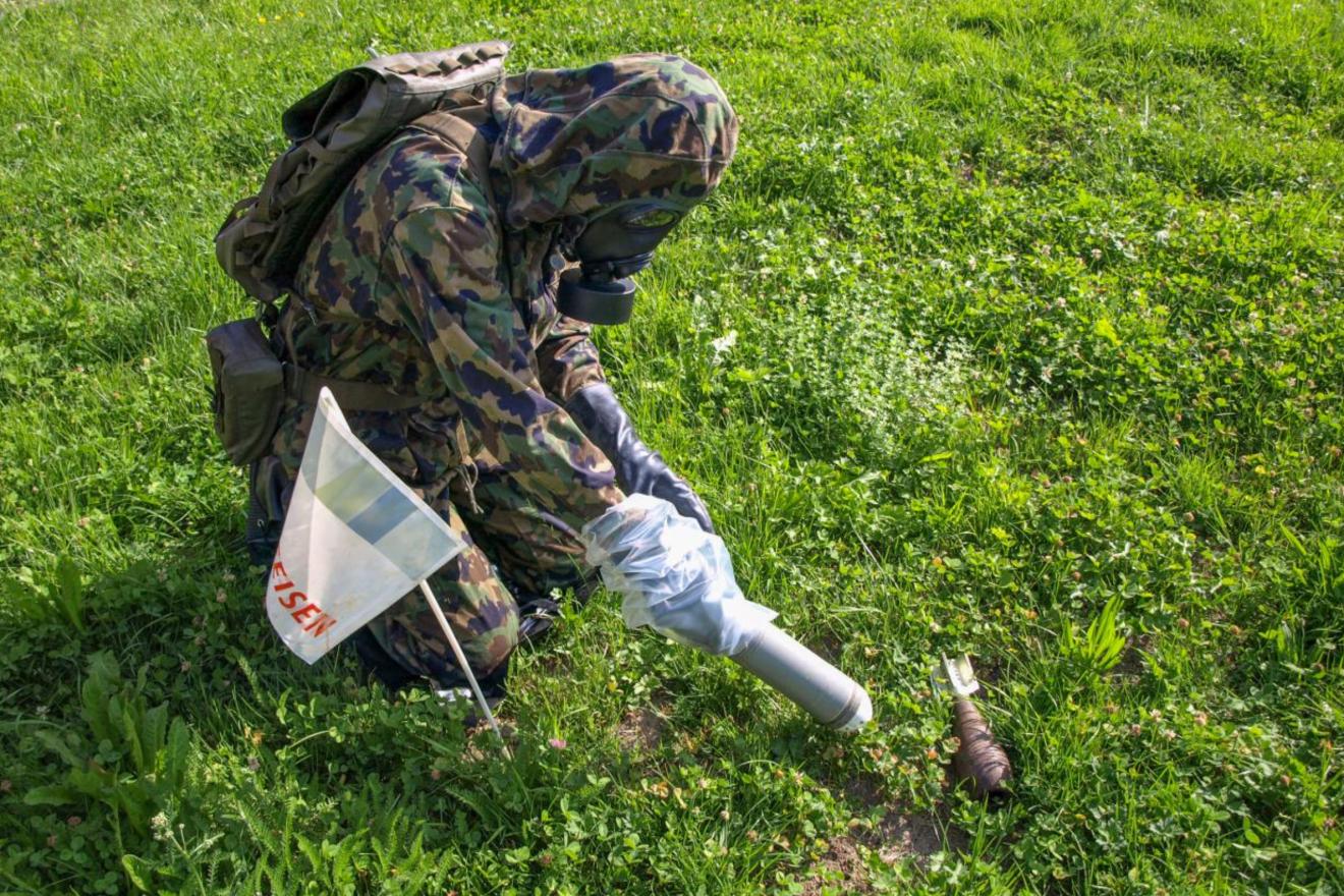 A photograph of a kneeling person in camouflage clothing and a gas mask in a field, disposing of a remnant of chemical warfare