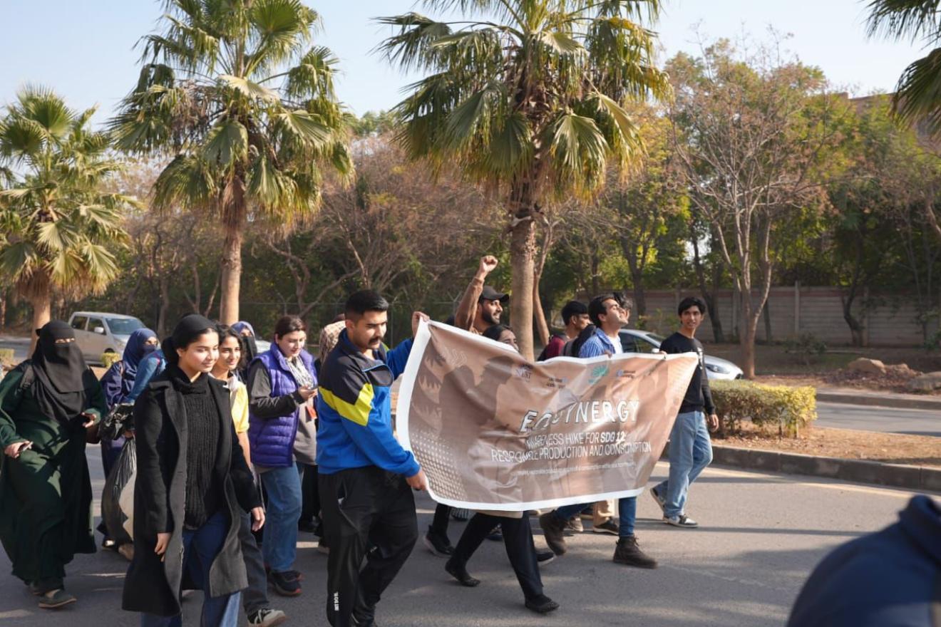 Group of students and staff walking together on a campus road holding an EcoSynergy SDG 12 banner.