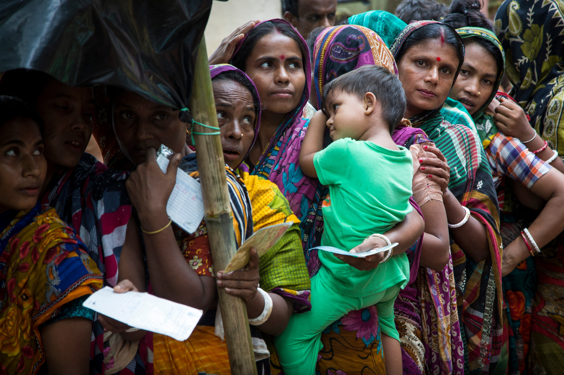 Several Rohingya women, one of whom is holding a child in her arms, wait in line.